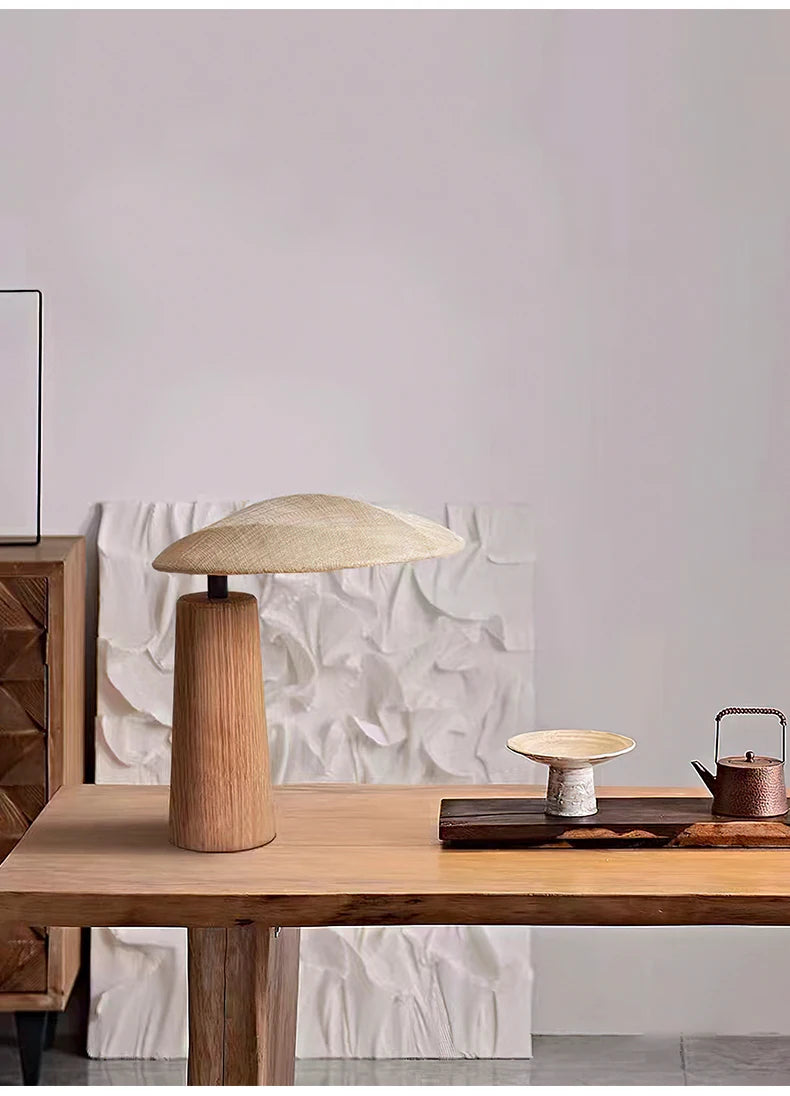 A Wabi Sabi inspired solid wood desk lamp with a natural finish, displaying an unconventional shape, resting on a wooden table with a textured wall in the background.